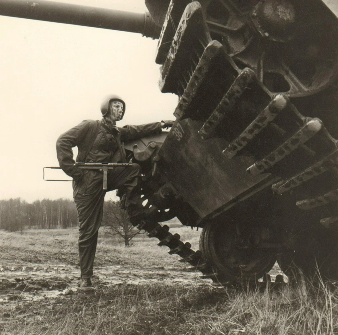 A person in a helmet and uniform stands beside a large tank, resting one foot on the tank's track in an open field with trees in the background. The image is black and white.