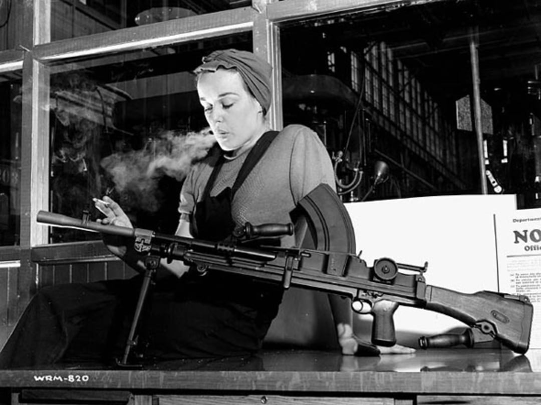 A woman in work clothes and a headscarf sits indoors, smoking a cigarette. She is assembling or inspecting a large machine gun on a workbench, with industrial equipment visible in the background.