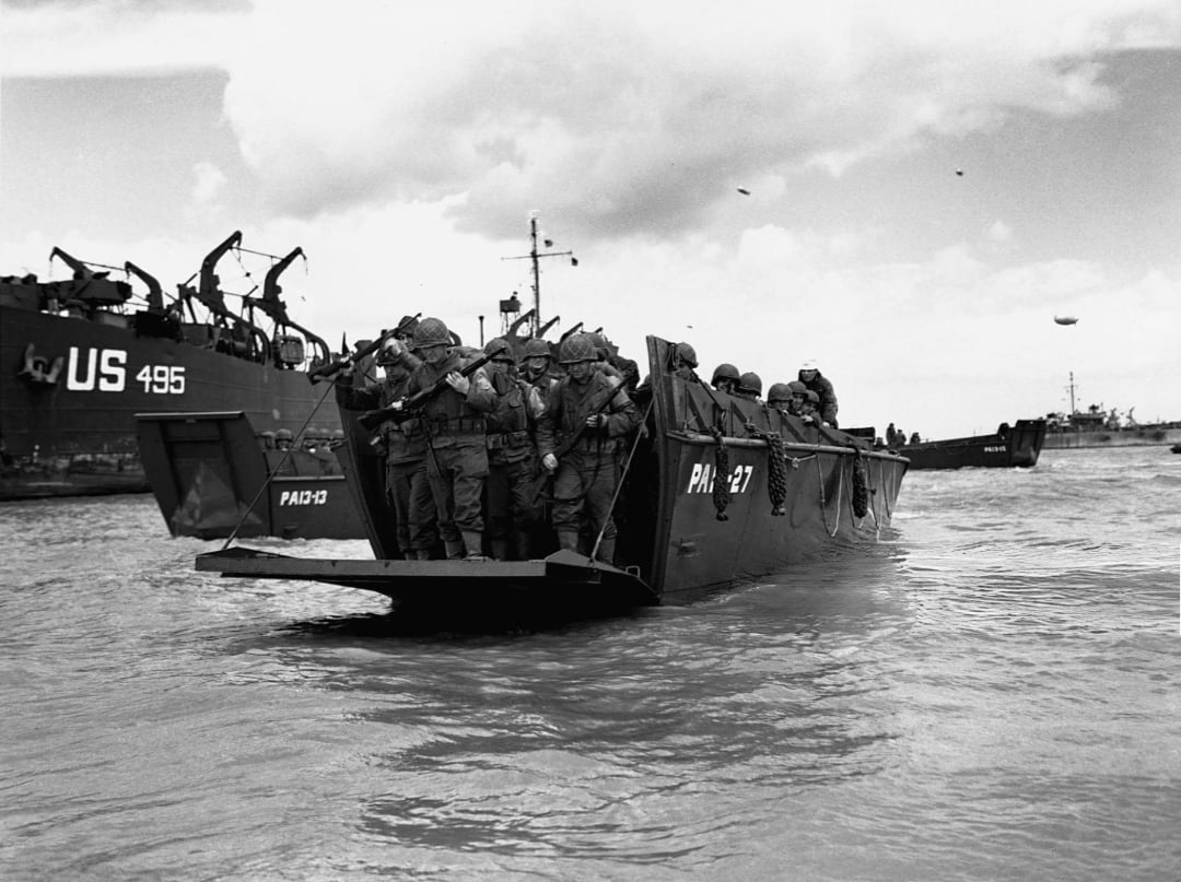 A group of soldiers in full gear stand crowded on a military landing craft approaching a beach, with larger ships visible in the background under a cloudy sky.