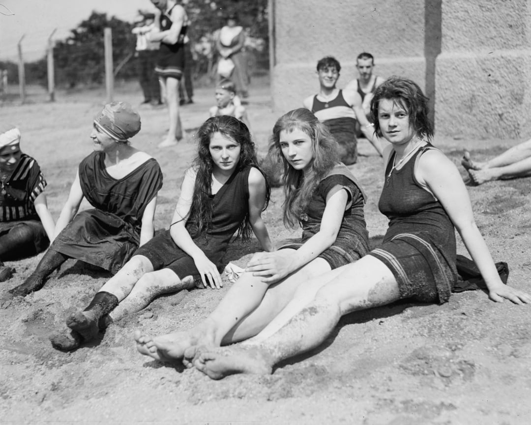 A group of young women and men in vintage swimsuits sit on sandy ground at a beach or lakeside, posing and relaxing in the sun, likely in the early 20th century. Some look at the camera, others face away.