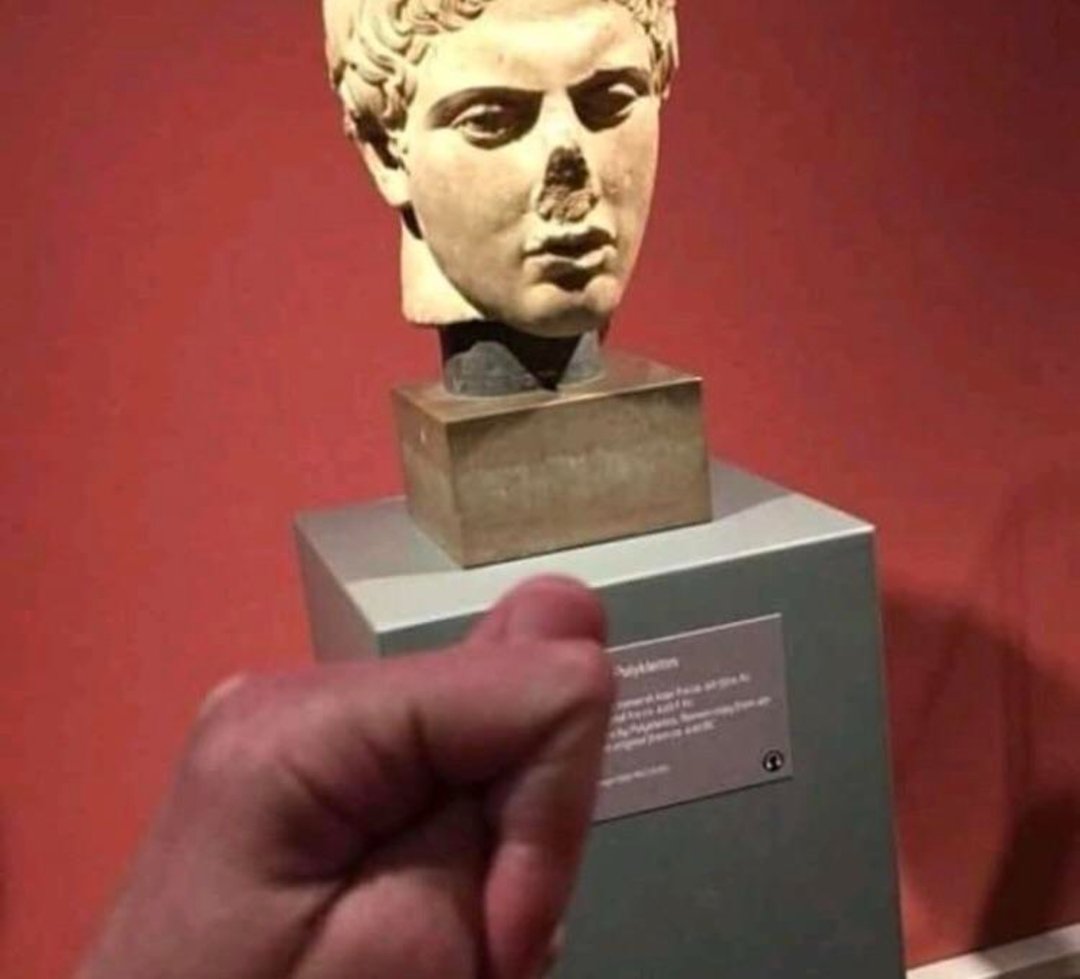 A person holds their thumb up in front of a broken ancient statue, aligning it so the thumb appears to replace the statue’s missing nose. The background is a museum display with a red wall.