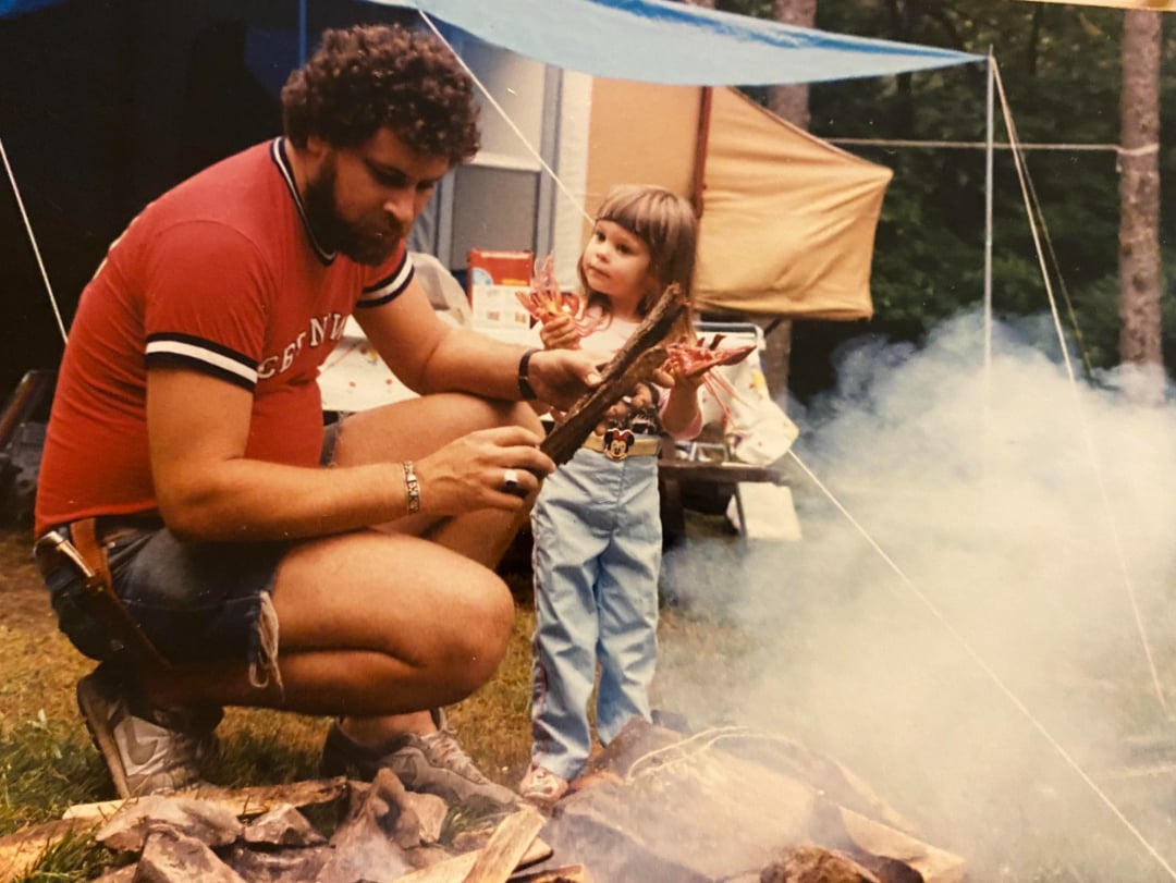 A man in a red shirt and shorts kneels by a campfire adding wood, while a young girl in jeans and a t-shirt stands beside him, looking at the fire. Tents and camping gear are visible in the background.