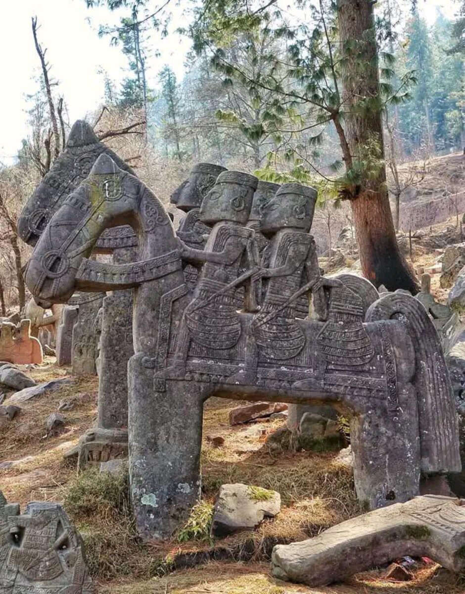 A large stone sculpture of three armored figures riding a horse stands outdoors among rocks and trees, surrounded by other carved stone monuments in a forested area.