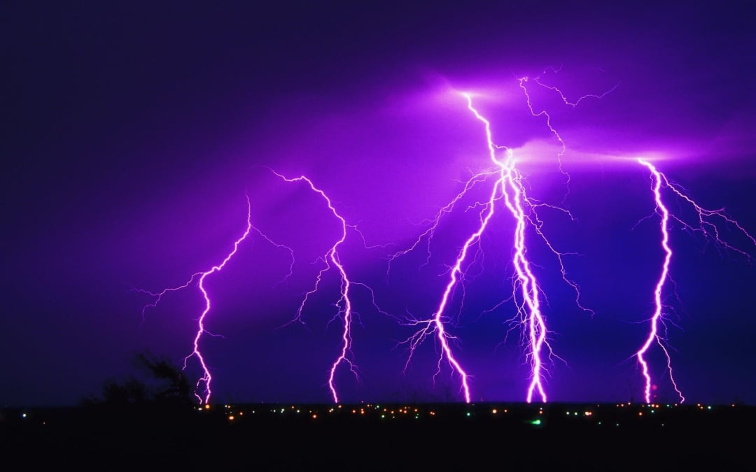 Multiple bright purple lightning bolts strike down from a dark, cloudy sky over a distant cityscape with small, colorful lights scattered across the horizon.
