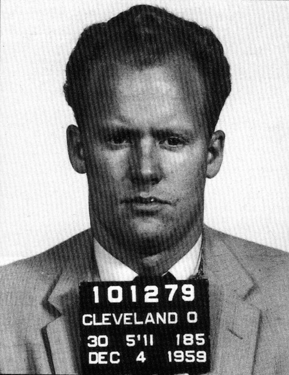 Black and white mugshot of a man wearing a suit. He holds a sign with the numbers 101279 and text reading "Cleveland O, 5'11, 185, Dec 4, 1959.
