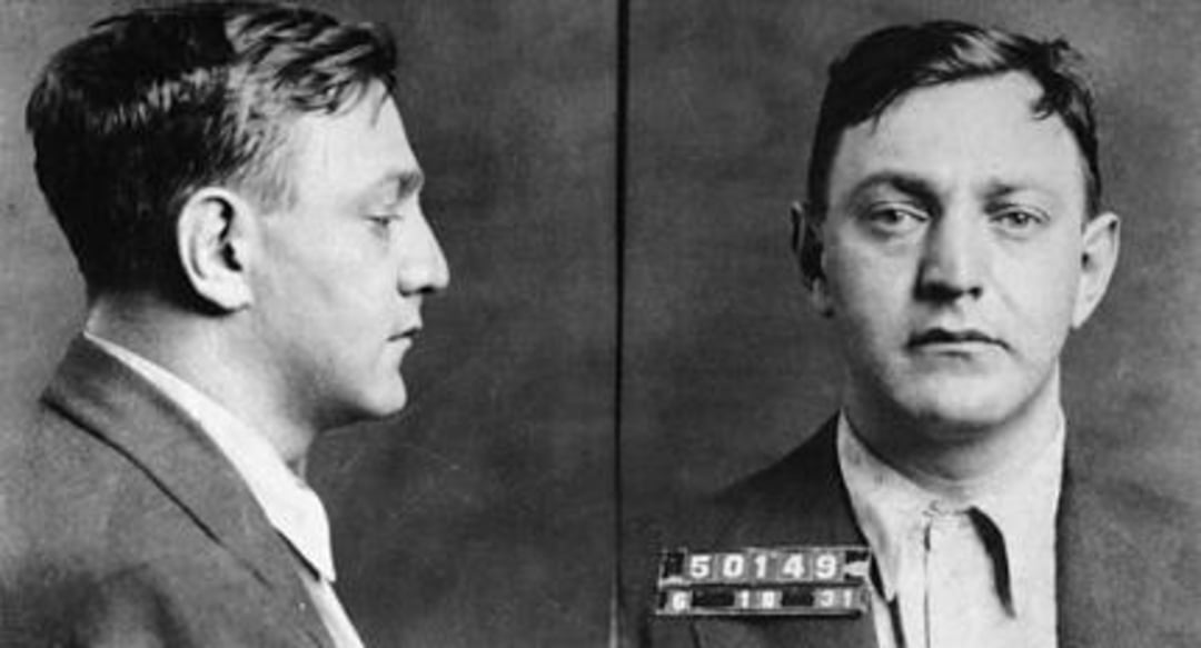 Black and white mugshot of a man in a suit, shown in both profile and front-facing views, with a neutral expression and an identification number "50149" displayed on his chest.