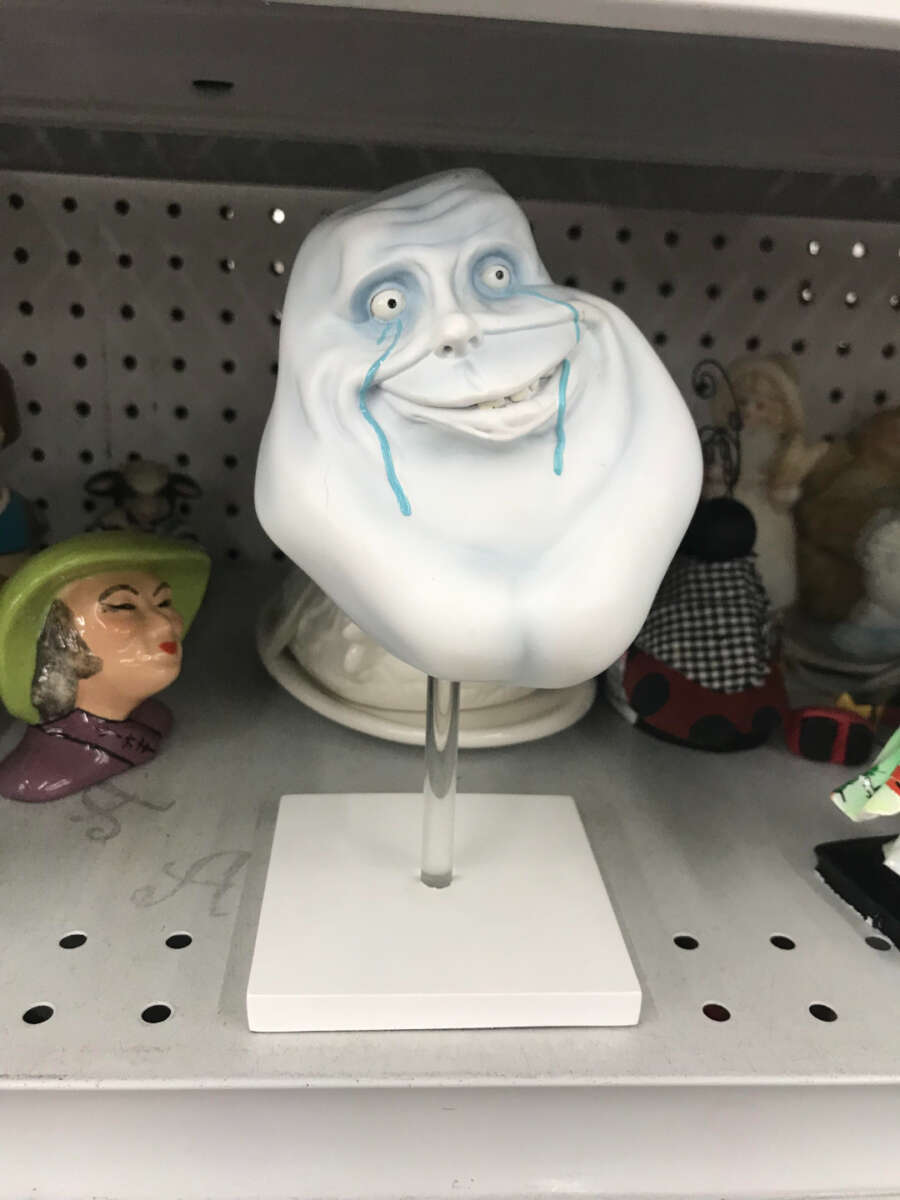 A white sculpture of a distorted, smiling face with exaggerated features and blue tears, displayed on a stand in a thrift store shelf among various ceramic figurines.