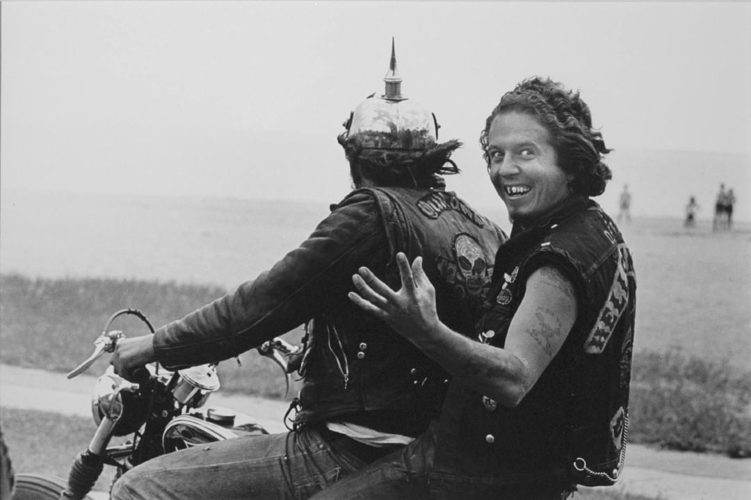 Two people ride a motorcycle; the driver wears a horned helmet and a vest with a skull patch, while the passenger looks back, grinning widely and gesturing animatedly with his hand. A beach and water are visible in the background.