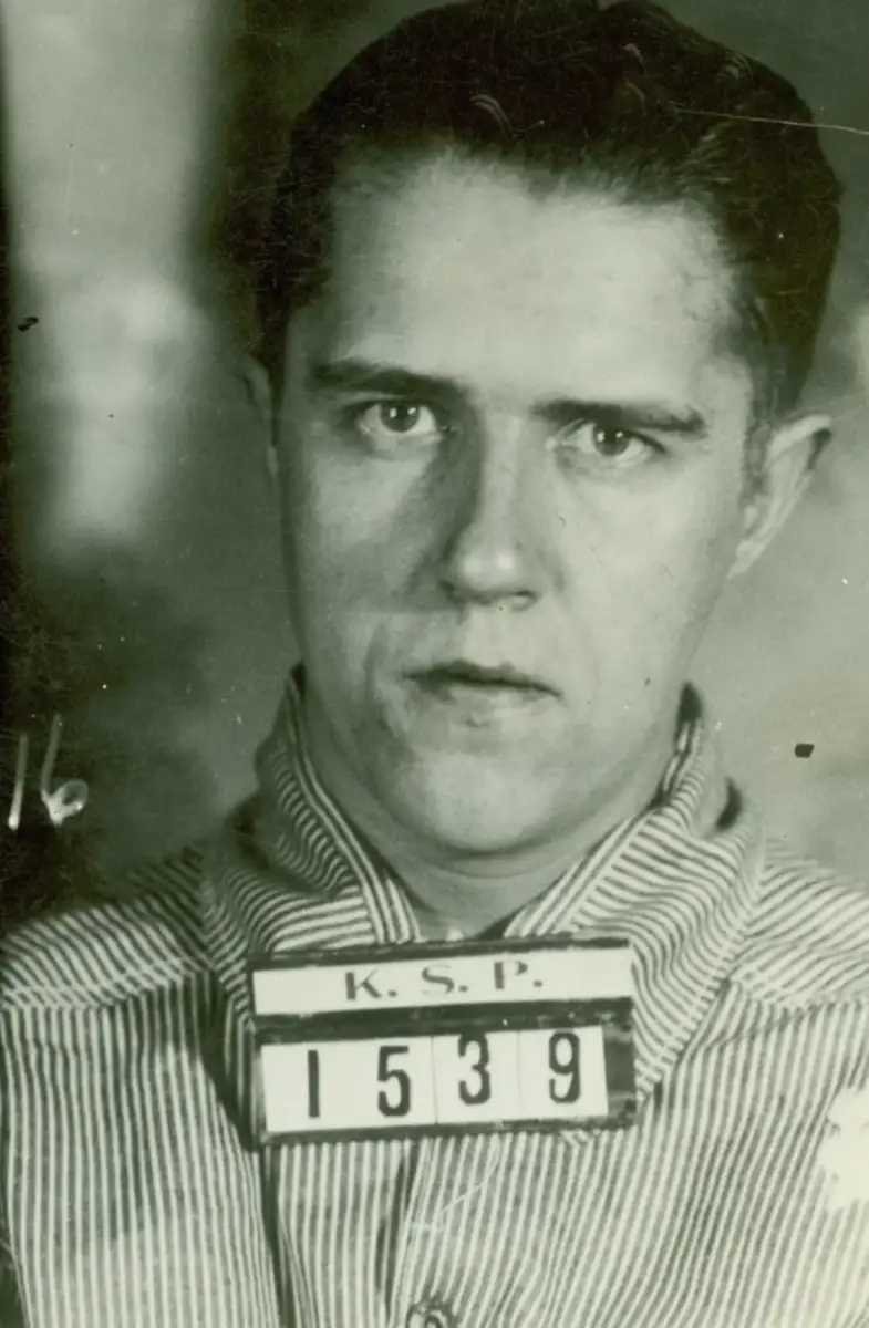 Black-and-white mugshot of a young man wearing a striped prison uniform, facing the camera. He holds a plaque labeled "K.S.P." with the number 1539. The background is blurred.