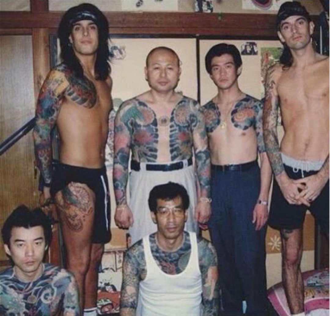 Six men, most shirtless, display extensive traditional Japanese tattoos while posing together indoors. The group stands and sits in a room with wooden walls and sliding doors.