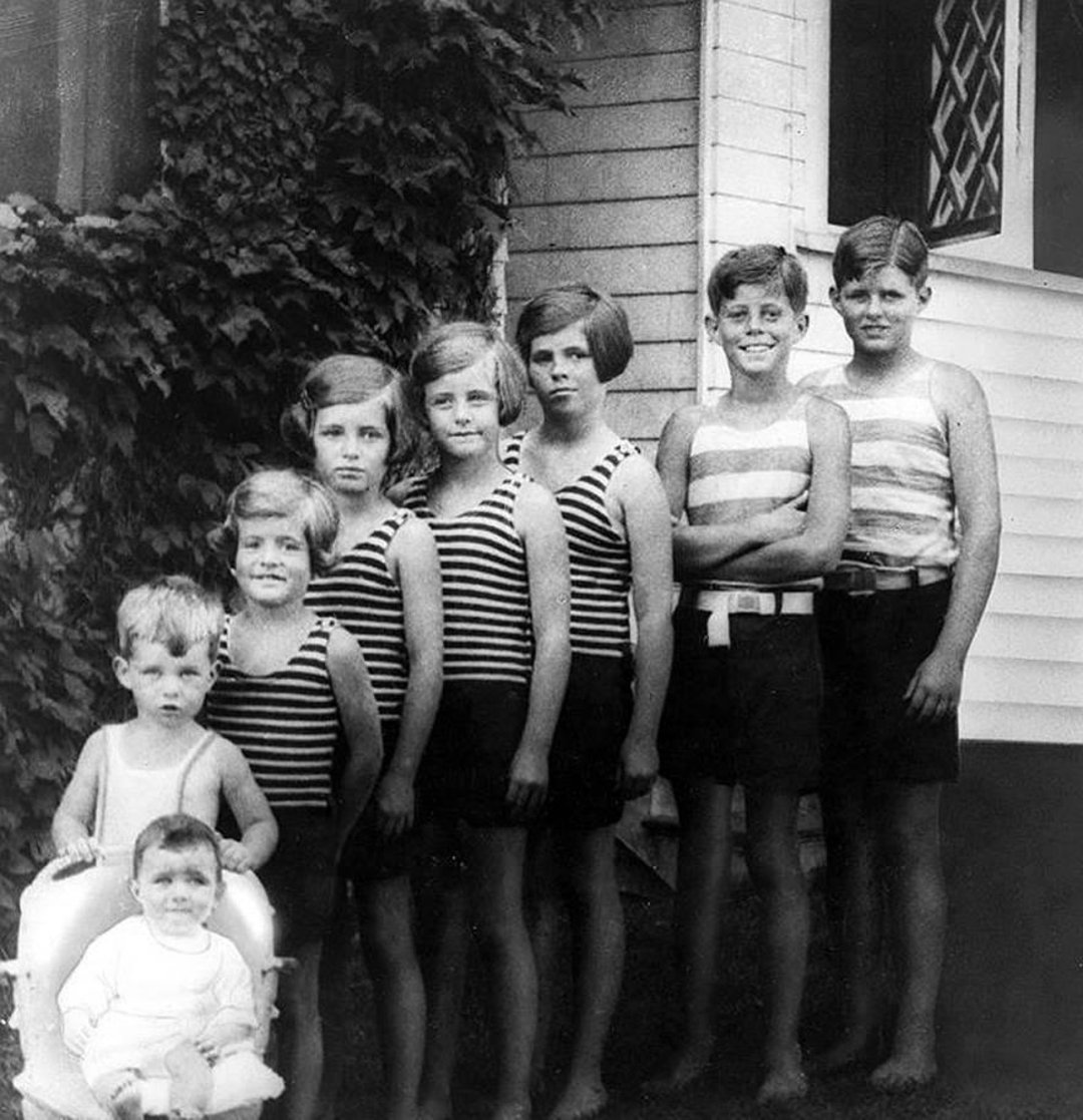 A vintage black-and-white photo of seven children standing in striped swimwear, with two younger children seated in front. They are outdoors next to a house partially covered in ivy.