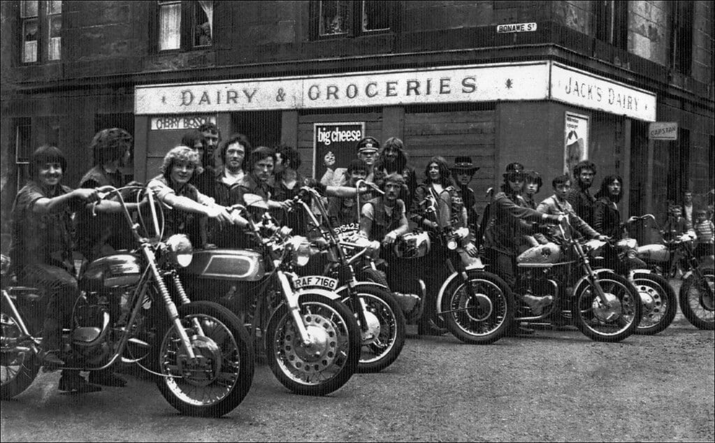 A group of people sit on motorcycles lined up in front of a corner store with a sign that reads "Dairy & Groceries" and "Jack’s Dairy." The image appears to be vintage, in black and white.
