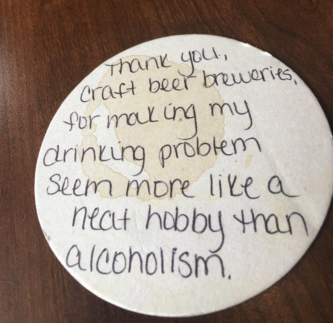 A round drink coaster with a coffee ring stain on a wooden table. Handwritten text says, “Thank you, craft beer breweries, for making my drinking problem seem more like a neat hobby than alcoholism.”