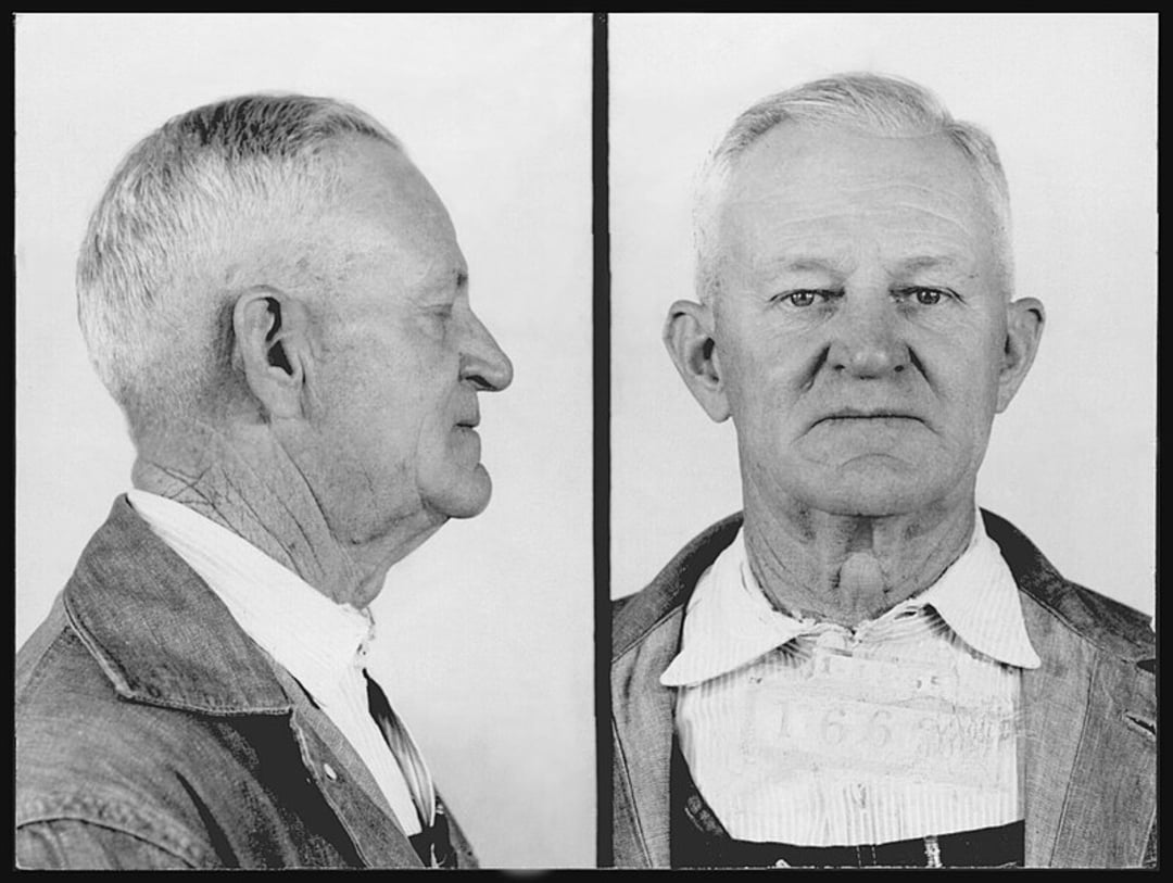 Black-and-white mugshot of an older man with short gray hair, shown in both side and front profiles, wearing a collared shirt and jacket. A card with numbers hangs around his neck.