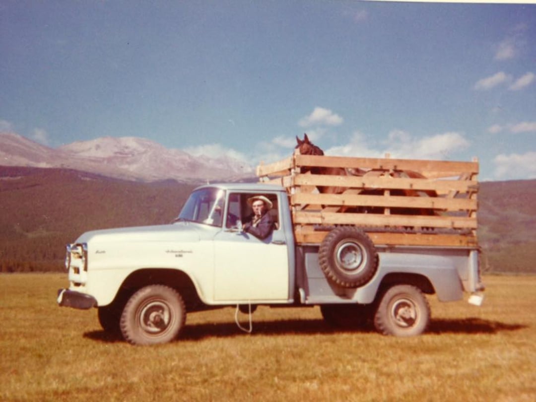 A vintage pickup truck with wooden side rails is parked on a grassy field; a person wearing a hat sits in the driver’s seat and a horse stands in the truck bed. Mountains and blue sky are in the background.