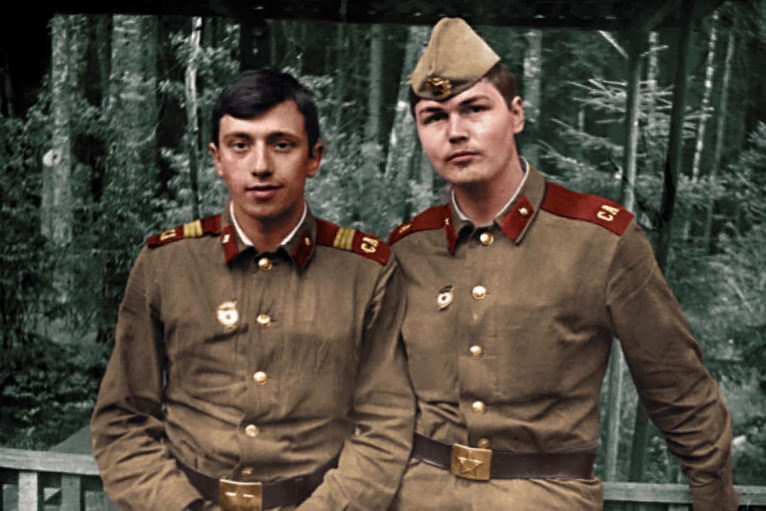 Two young men in vintage military uniforms with red accents stand side by side outdoors, in front of a forested background. One wears a cap; both have badges on their jackets and serious expressions.