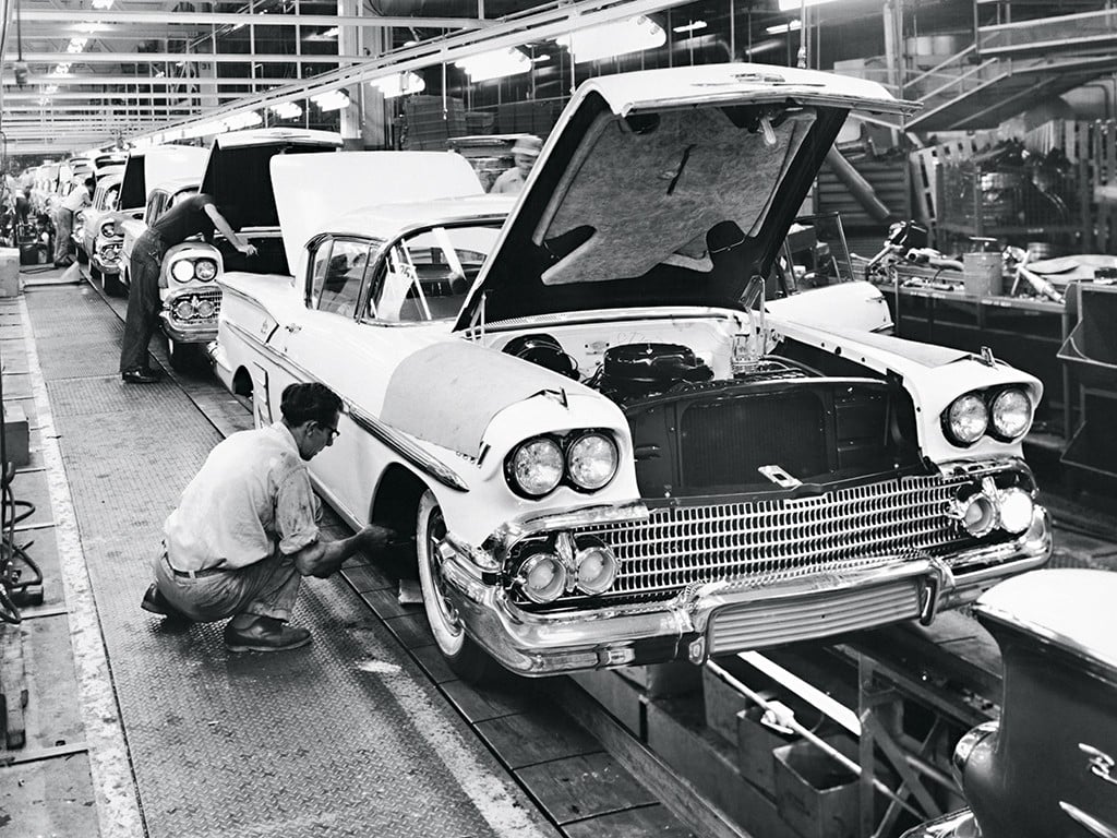 Black-and-white photo of workers assembling classic cars on an automotive factory production line, with car hoods open and one worker crouched by a front tire, focusing on vehicle details.