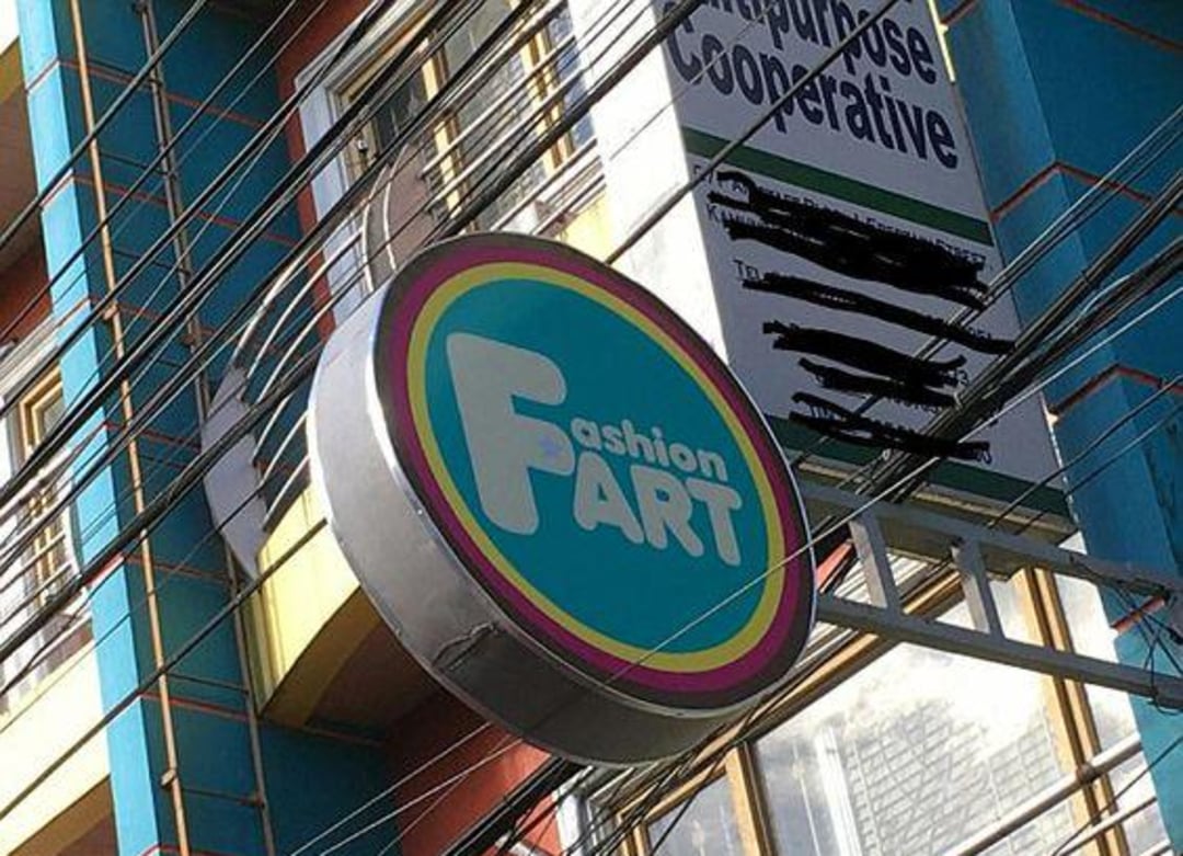 A circular store sign reads "Fashion Fart" in colorful letters, hanging on the side of a building with blue walls and yellow trim. Power lines crisscross in front of the sign.