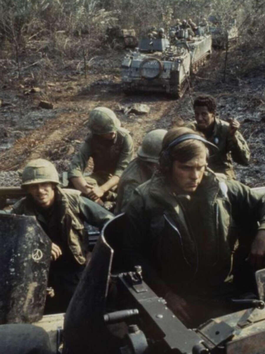 A group of soldiers in military gear sit and stand on armored vehicles in a muddy, forested area, with more soldiers and vehicles visible in the background. The scene appears tense and focused.