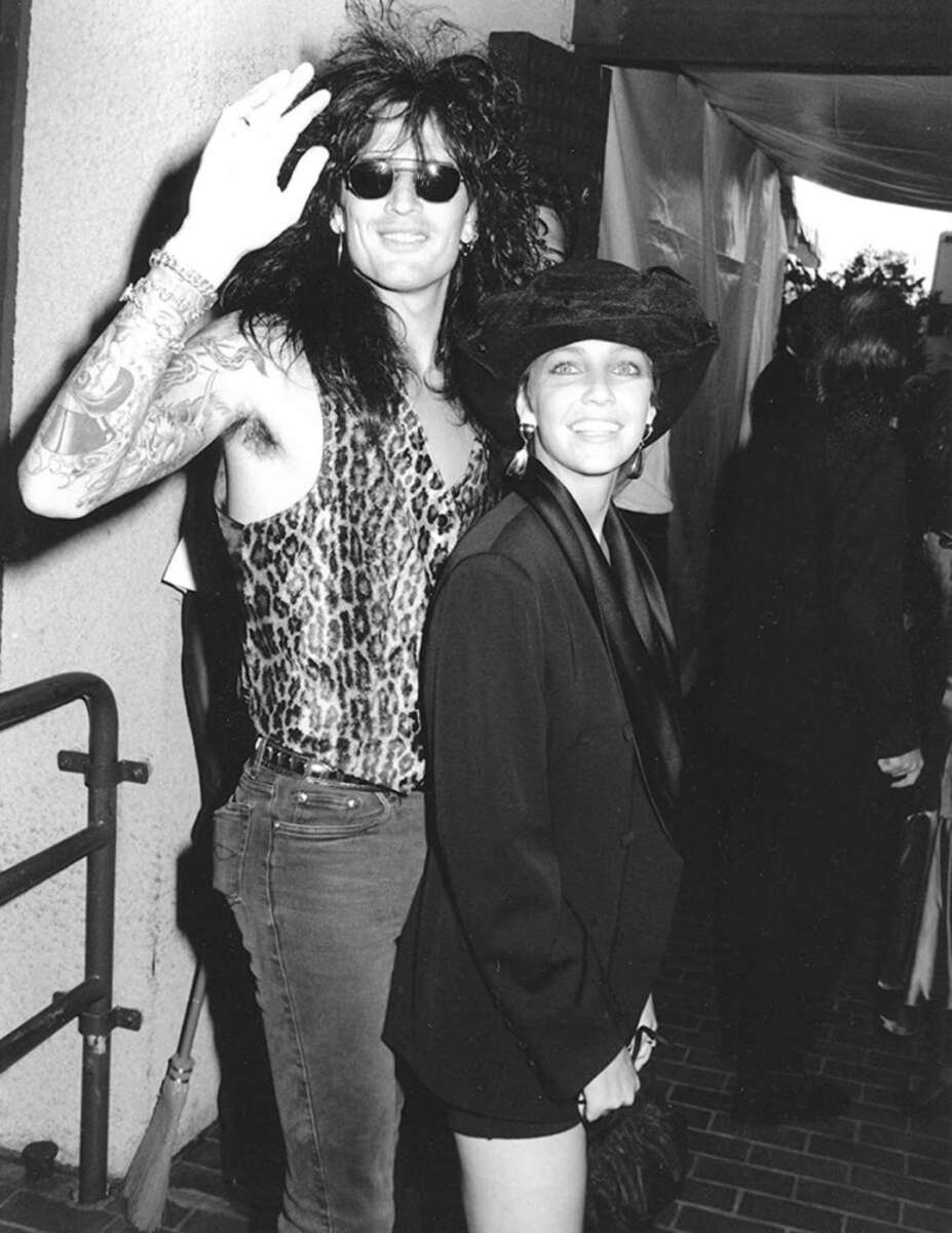 A man in a sleeveless leopard print shirt with long dark hair, sunglasses, and tattoos waves while standing next to a smiling woman in a black hat and blazer. The photo is black and white and appears to be at an event.