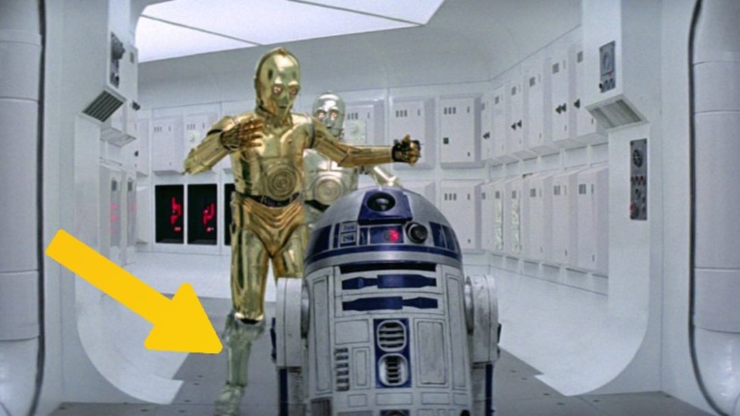 C-3PO and R2-D2, two droids from Star Wars, walk down a white spaceship corridor. A large yellow arrow points to C-3PO’s lower right leg, highlighting it for emphasis.
