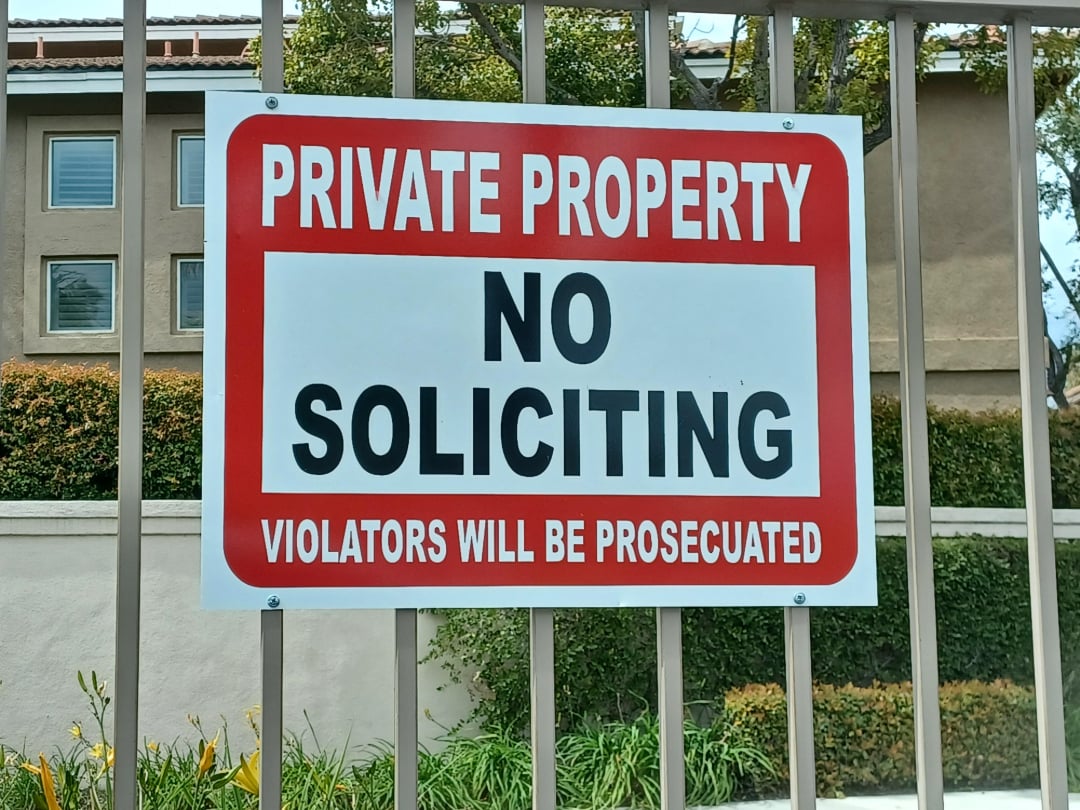 A sign on a fence reads "PRIVATE PROPERTY NO SOLICITING VIOLATORS WILL BE PROSECUTED" in bold letters, warning against solicitation. An apartment building and greenery are visible in the background.