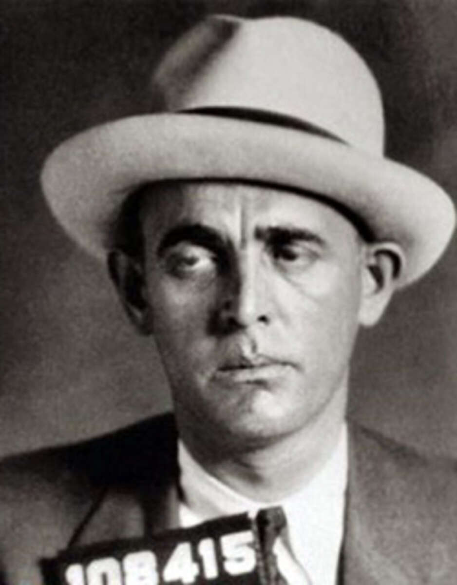 Black and white photo of a man wearing a light-colored hat and suit, looking to the side with a serious expression. He is holding a sign with the number "108415.