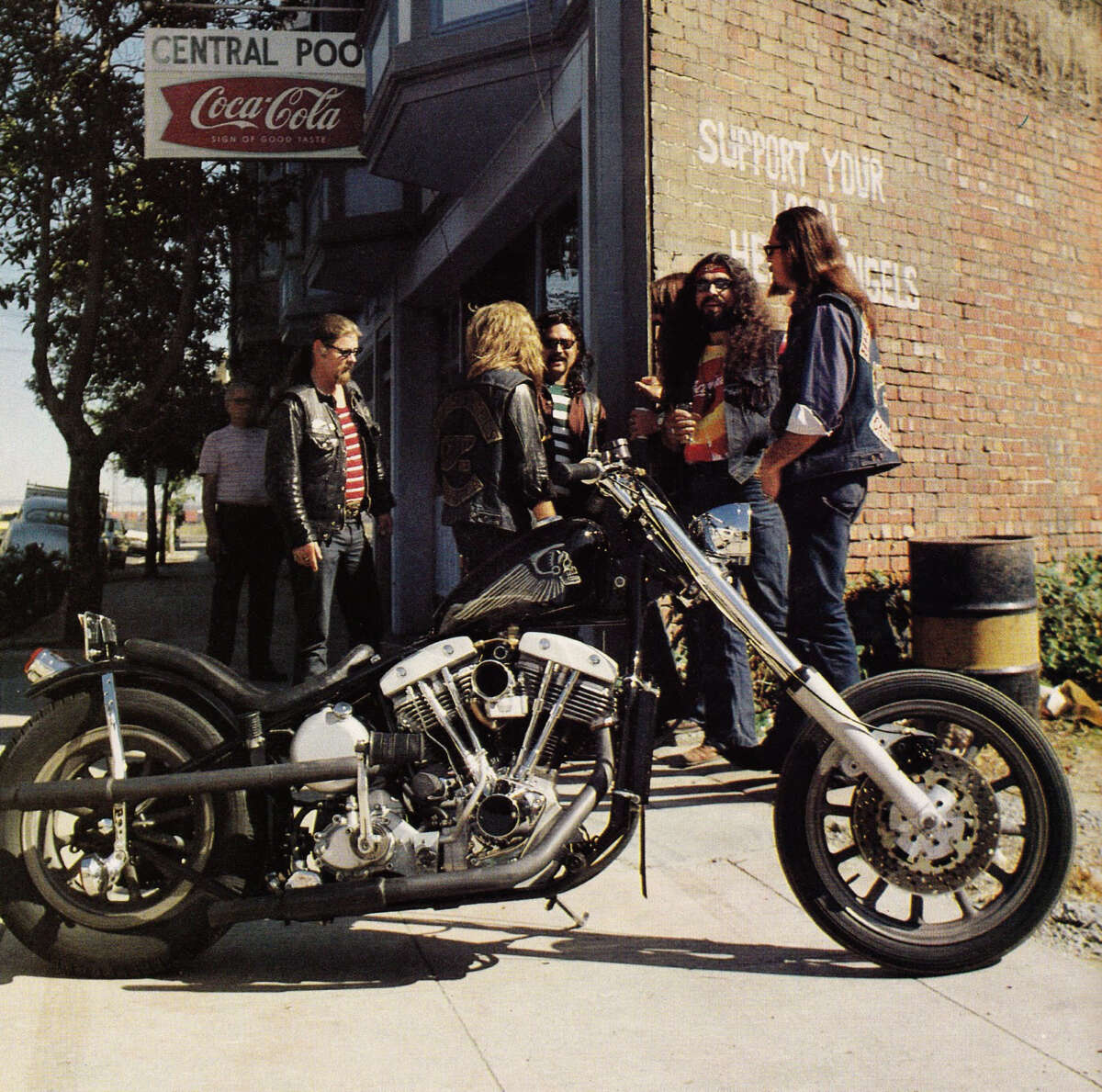 A group of people in leather jackets gather and talk near a brick wall and a custom chopper motorcycle parked on the sidewalk outside a building with a "Coca-Cola" sign.