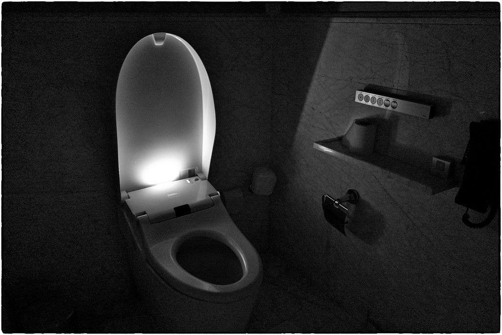 A modern toilet with the lid open is softly illuminated in a dark bathroom. Control buttons, a toilet paper holder, and a small cup are mounted on the wall nearby.