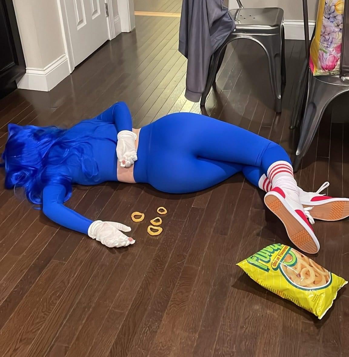 A person in a blue Sonic the Hedgehog costume lies face-down on a hardwood floor with white gloves, red shoes, and rings made from onion snacks beside them. An open bag of Funyuns is spilled on the floor.