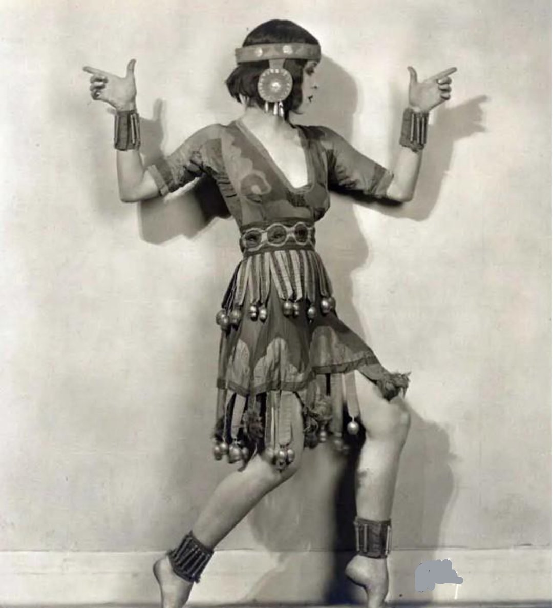 A woman in a costume strikes a dramatic pose, arms bent and pointed outward, wearing a headband, round earrings, bracelets, and a dress with decorative tassels, against a plain background.