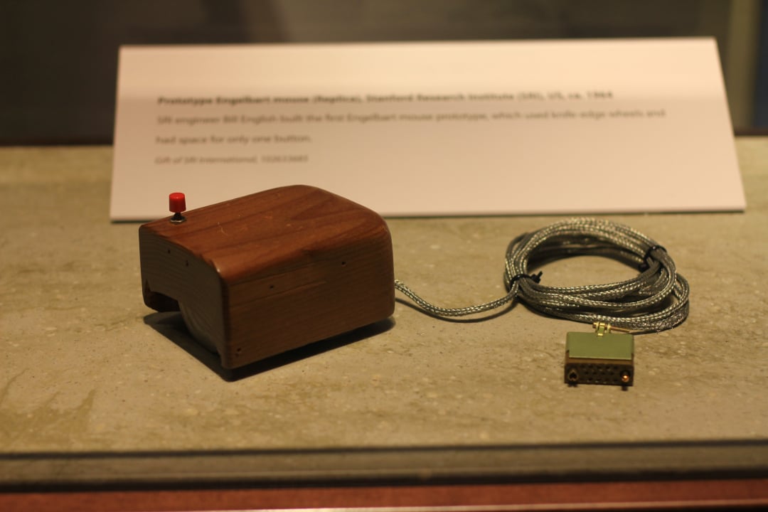 A wooden prototype computer mouse with a single red button and a cord attached, displayed on a gray surface with an informational plaque in the background.