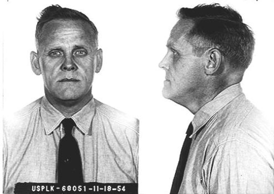 Black and white mugshot of a middle-aged man with short hair, wearing a buttoned shirt and dark tie, shown facing forward and in profile. A placard below displays "USPLK-68051-11-18-54.
