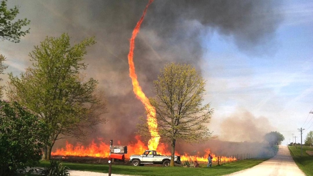 A large fire whirl, or “fire tornado,” rises from burning grass near a road where a white pickup truck and two people stand nearby. Thick smoke fills the sky amid green trees and a rural landscape.