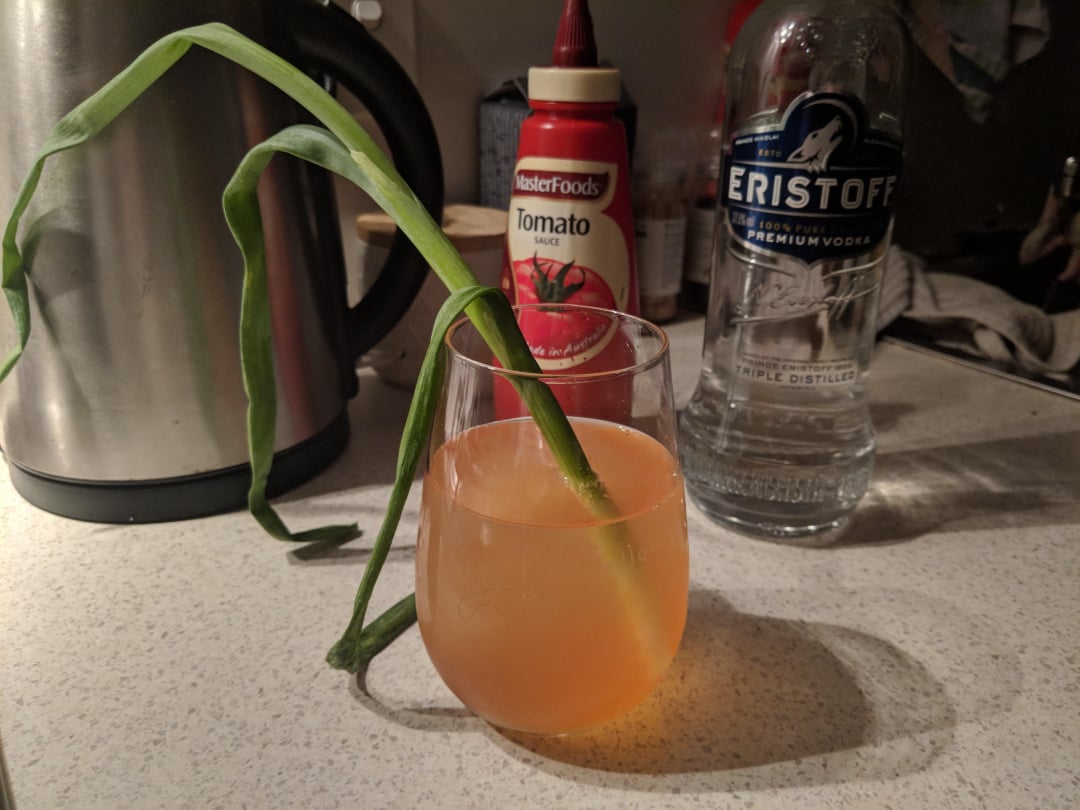 A cocktail in a glass with a long green stalk as garnish sits on a kitchen counter, next to a bottle of vodka, tomato sauce, and an electric kettle.