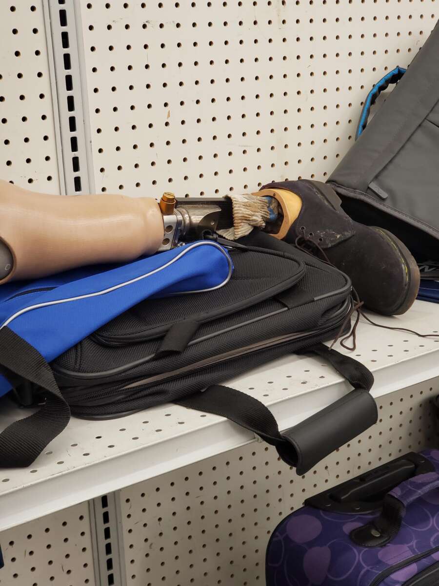 A prosthetic leg wearing a brown boot rests on top of a black and blue laptop bag on a white store shelf with pegboard backing. Other bags are partially visible nearby.
