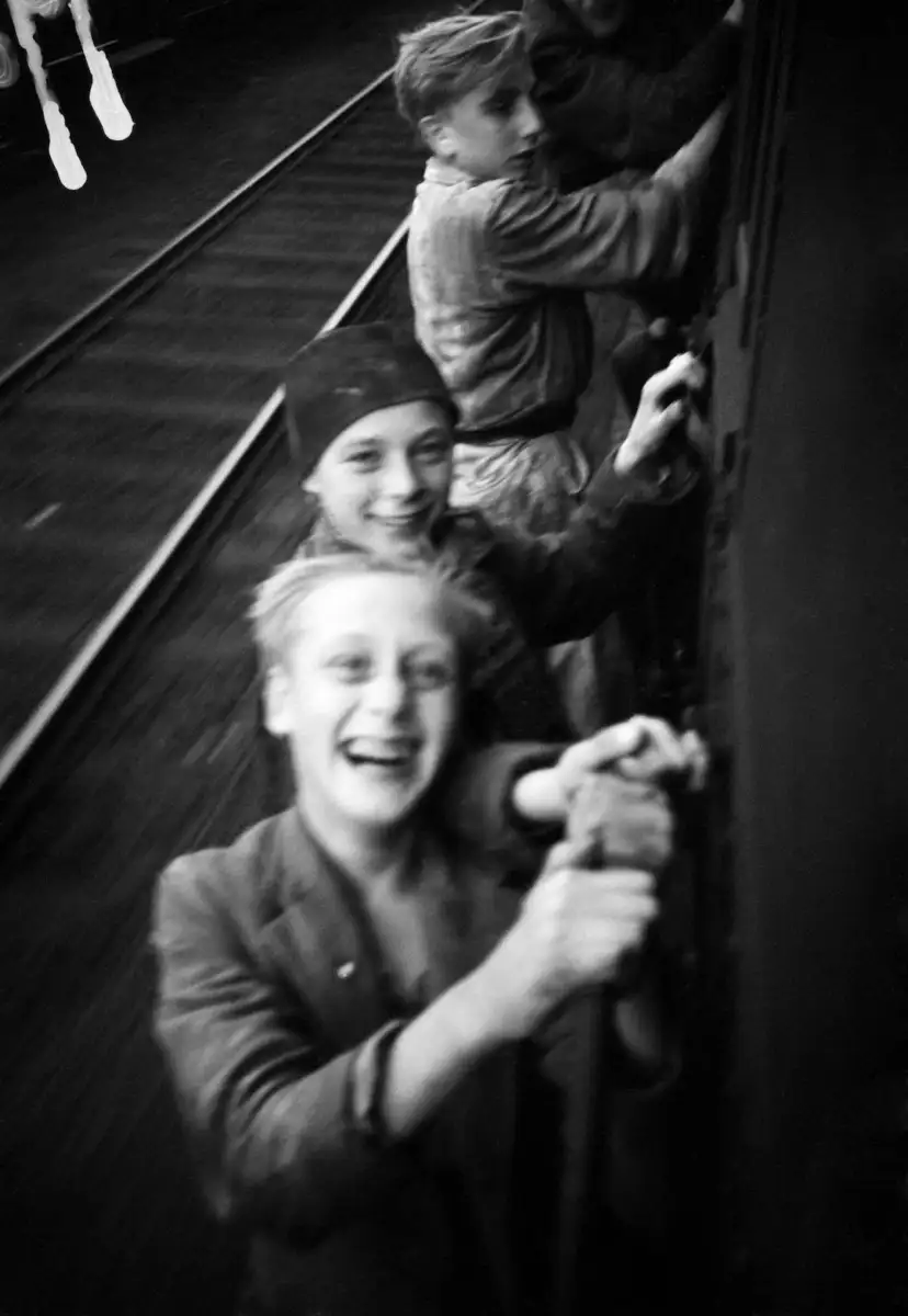 Three boys, smiling and laughing, hold onto the side of a moving train. The background shows blurred train tracks, emphasizing the motion. The image is in black and white.