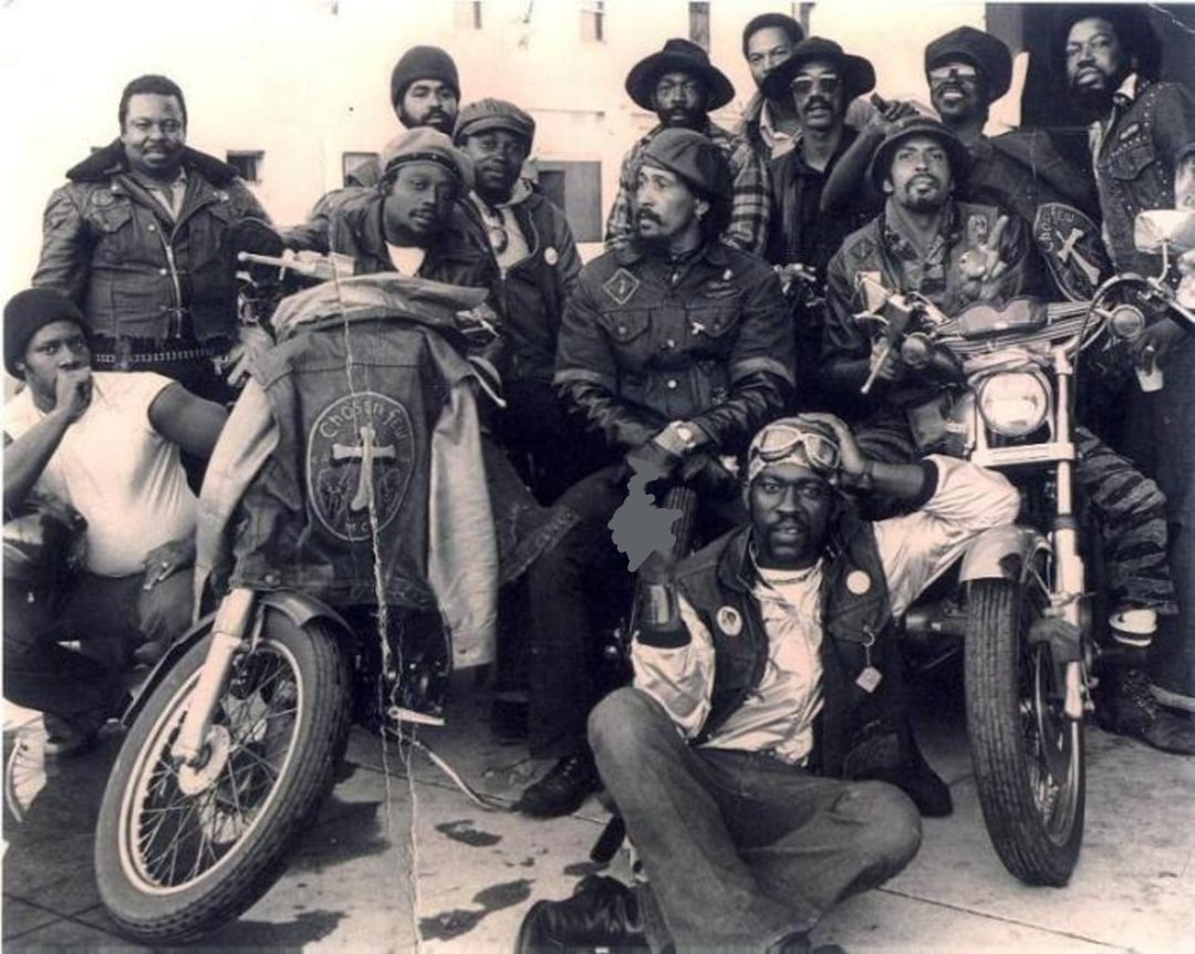 A group of Black men pose together with motorcycles, many wearing leather vests and jackets with patches. Some sit on bikes, others stand close, looking at the camera in a confident, friendly group pose.