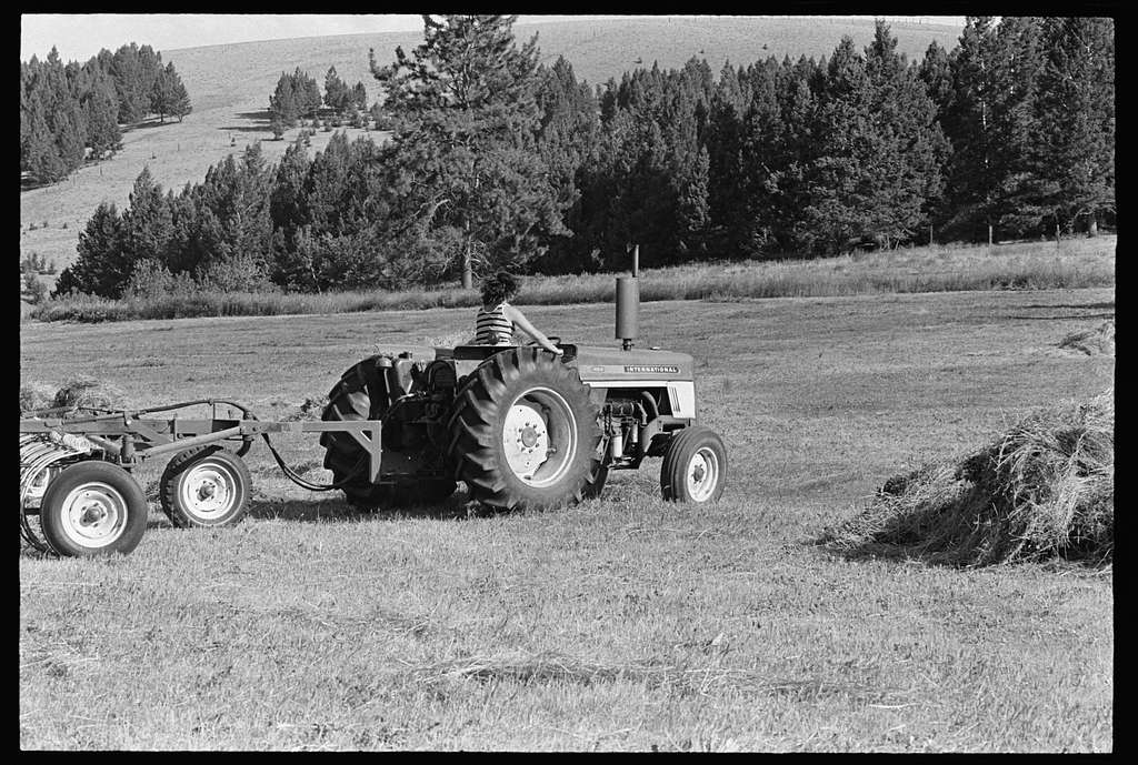 A tractor pulling farming equipment works in an open field with trees and hills in the background. There is a pile of hay nearby.