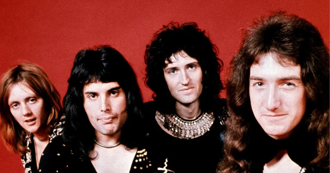 Four men with long hair stand close together against a solid red background, looking at the camera and smiling slightly. They are dressed in dark, patterned clothing with some metallic embellishments.