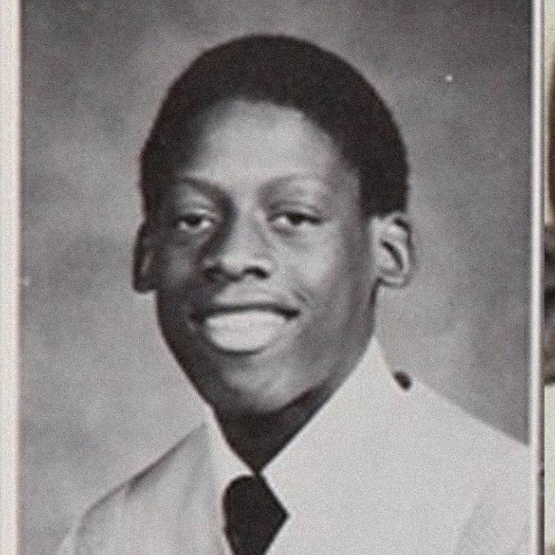 A black and white yearbook-style portrait of a young man smiling, wearing a light-colored dress shirt and dark tie, with short hair, pictured against a plain background.