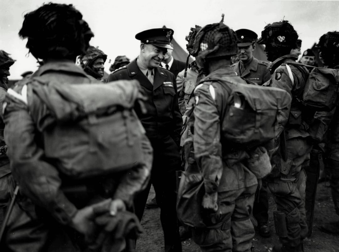 A military officer smiles and speaks with a group of soldiers in uniform, wearing backpacks and helmets, outdoors in a wartime setting.