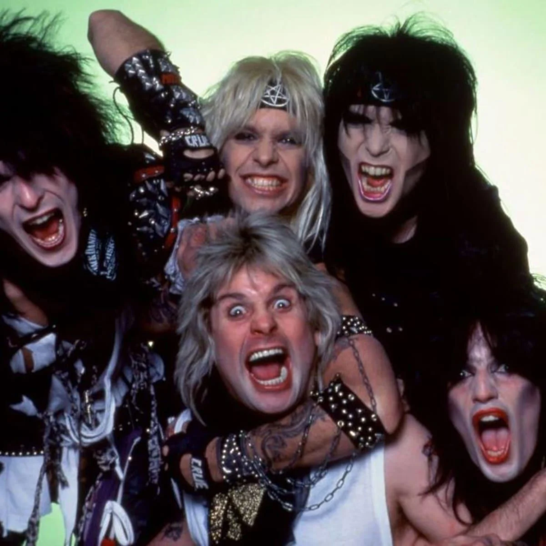 Five members of a glam rock band pose with wild expressions, open mouths, and exaggerated makeup, wearing spiked accessories and big, teased hair against a light green background.
