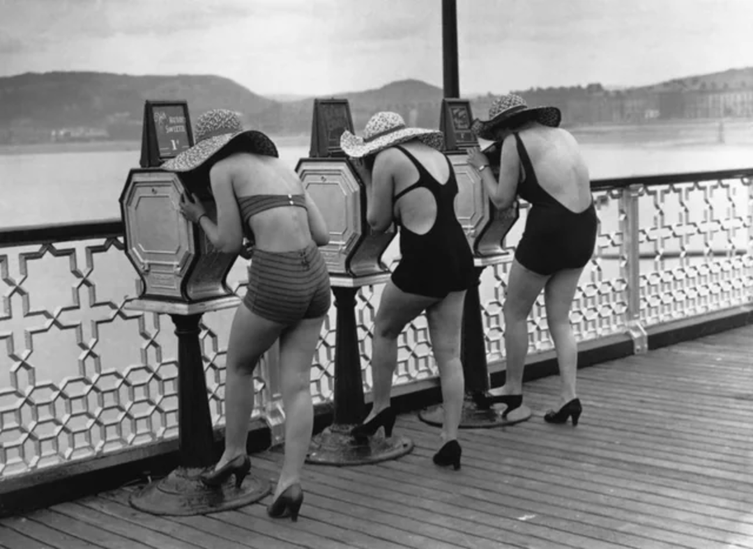 Three women in swimsuits, hats, and heels look into coin-operated viewing machines on a boardwalk, with water and hills visible in the background. The scene appears vintage or from the mid-20th century.
