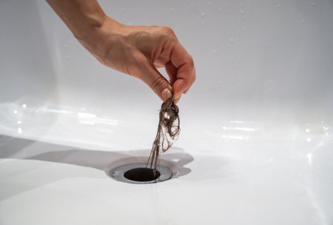 A hand is pulling a clump of hair out of a white sink drain, illustrating the process of cleaning a clogged drain.