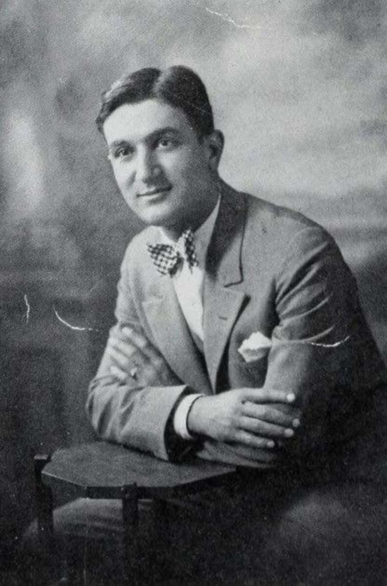 Black and white photo of a young man in a suit and bow tie, seated with arms crossed on a chair, looking at the camera and smiling slightly. The background is softly blurred.
