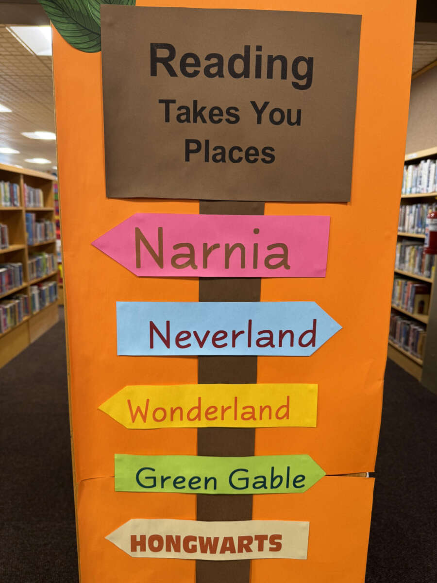 A library display with the sign “Reading Takes You Places” and colorful arrows pointing to fictional locations: Narnia, Neverland, Wonderland, Green Gable, and Hongwarts. Bookshelves are visible in the background.