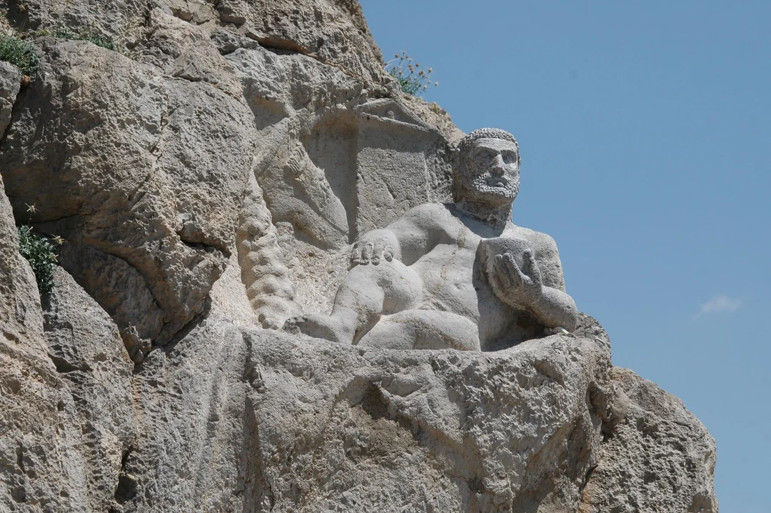 A large stone relief of a seated, bearded man is carved into a rocky cliff, holding an object in his left hand, with a clear blue sky in the background.
