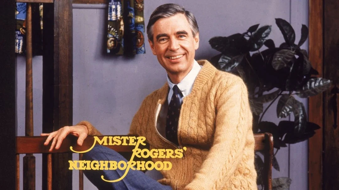 A man wearing a tan cardigan and tie sits on a bench, smiling warmly. Text on the image reads "Mister Rogers' Neighborhood." A plant and colorful window curtain are visible in the background.