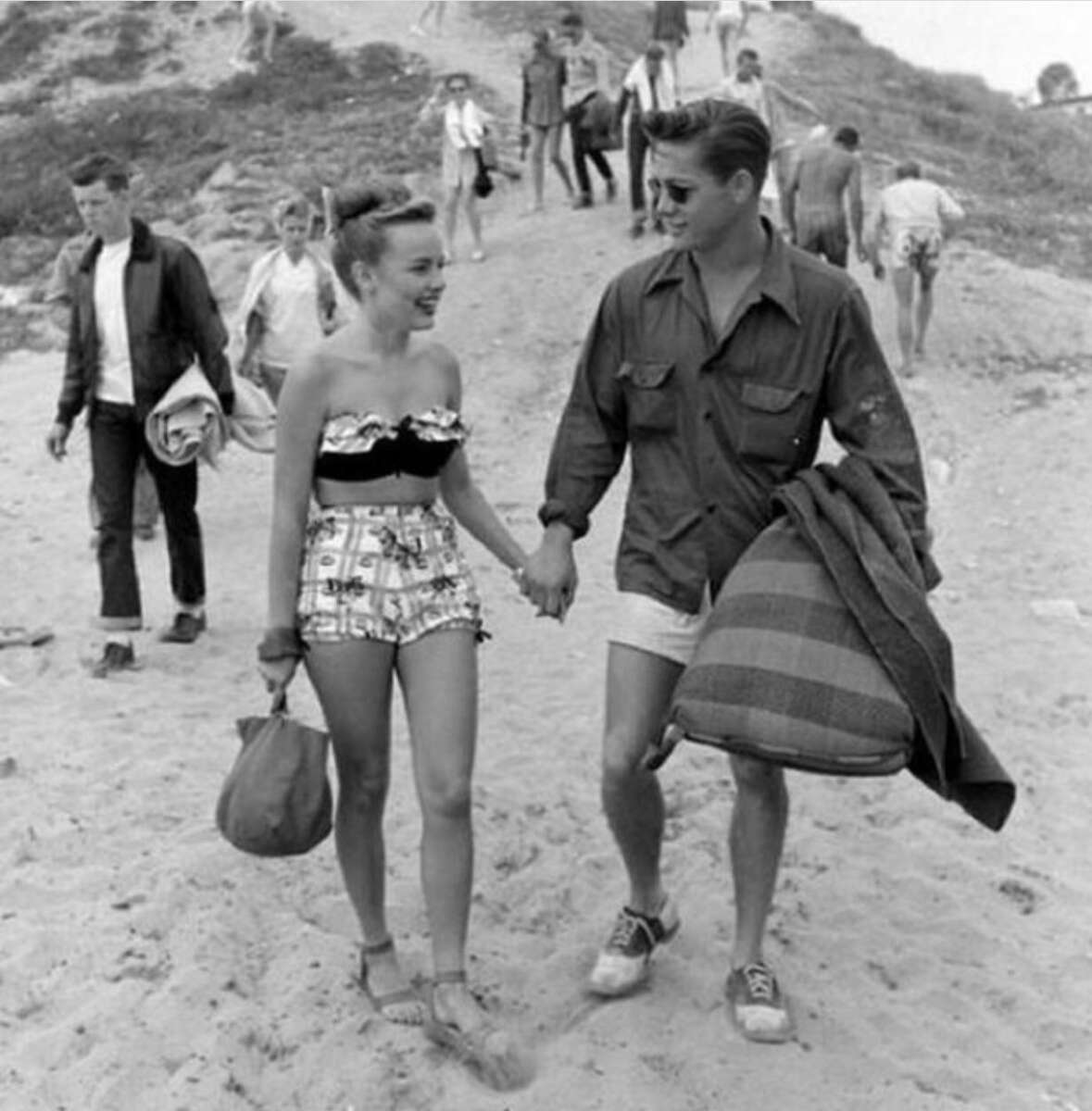 A young woman in a patterned swimsuit and shorts holds hands with a man in a shirt and shorts carrying a blanket, walking on a sandy beach with other people in the background.
